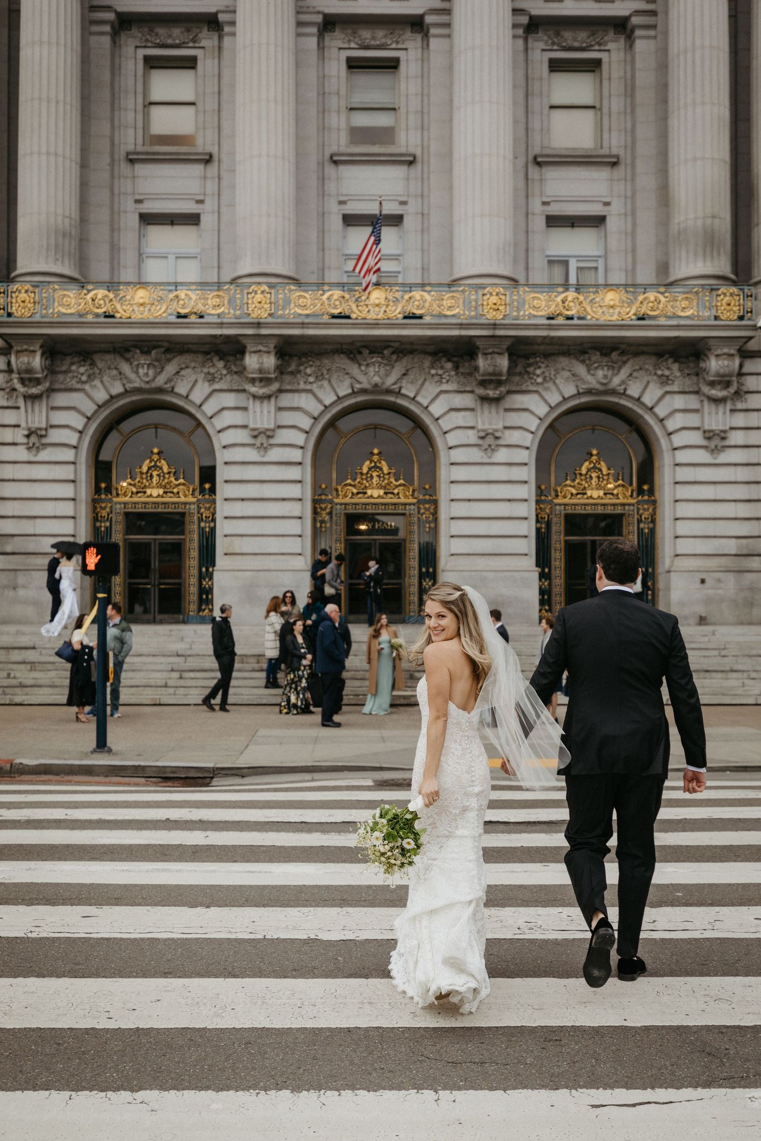 A wedding couple ascends grand stone steps of a historic building with ornate golden doorways and columns.