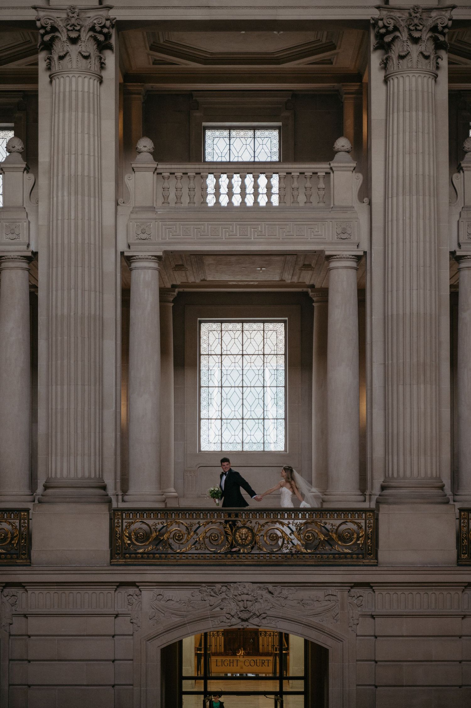 A wedding couple stands on an ornate balcony in a grand architectural setting.