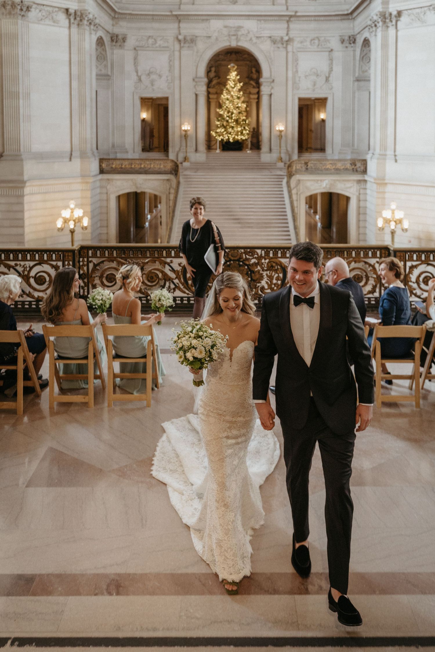 A wedding couple walks down marble stairs in an elegant venue with ornate architecture and a glowing Christmas tree.