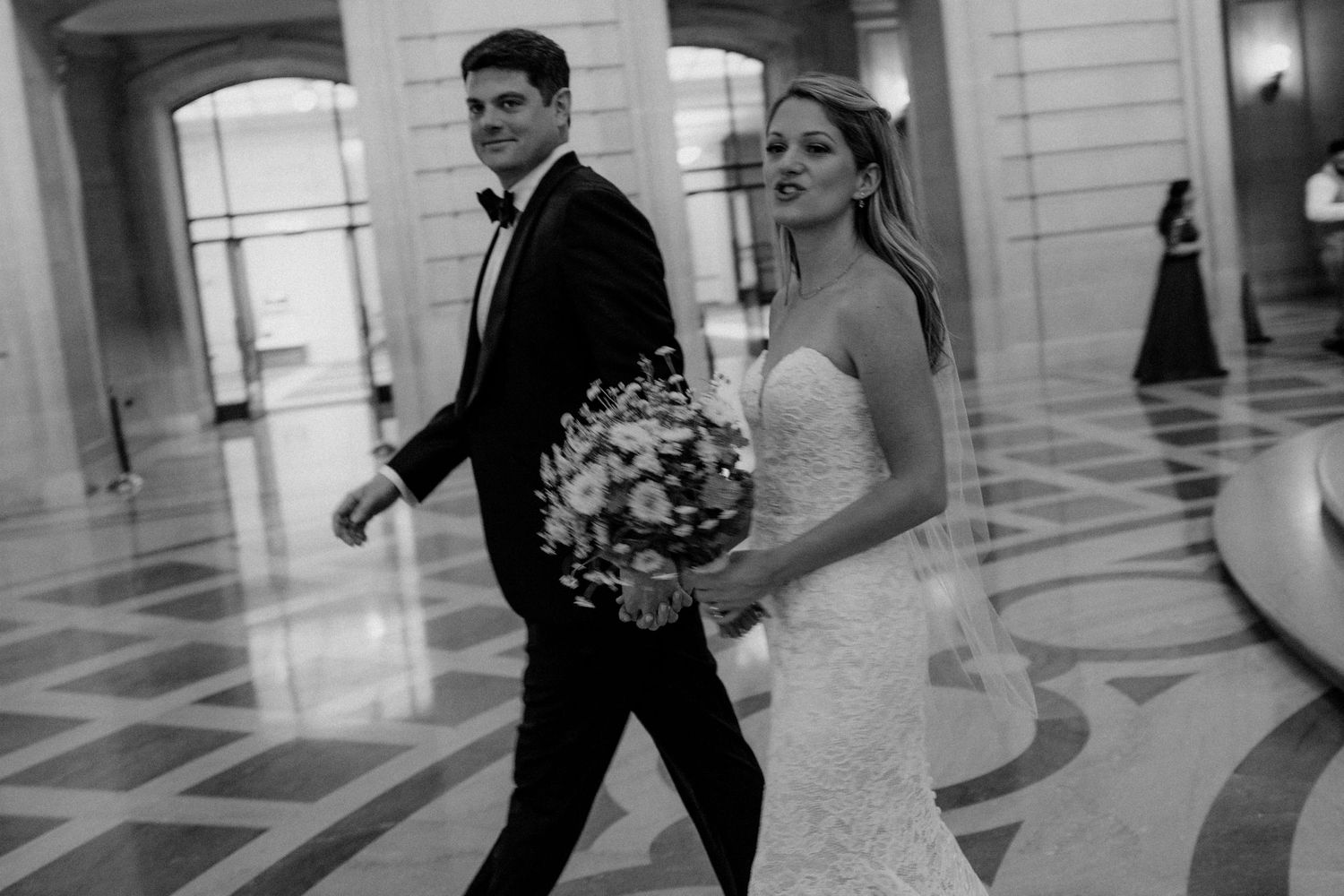 Elegant couple walking through marble hallway with bridal bouquet and formal attire.