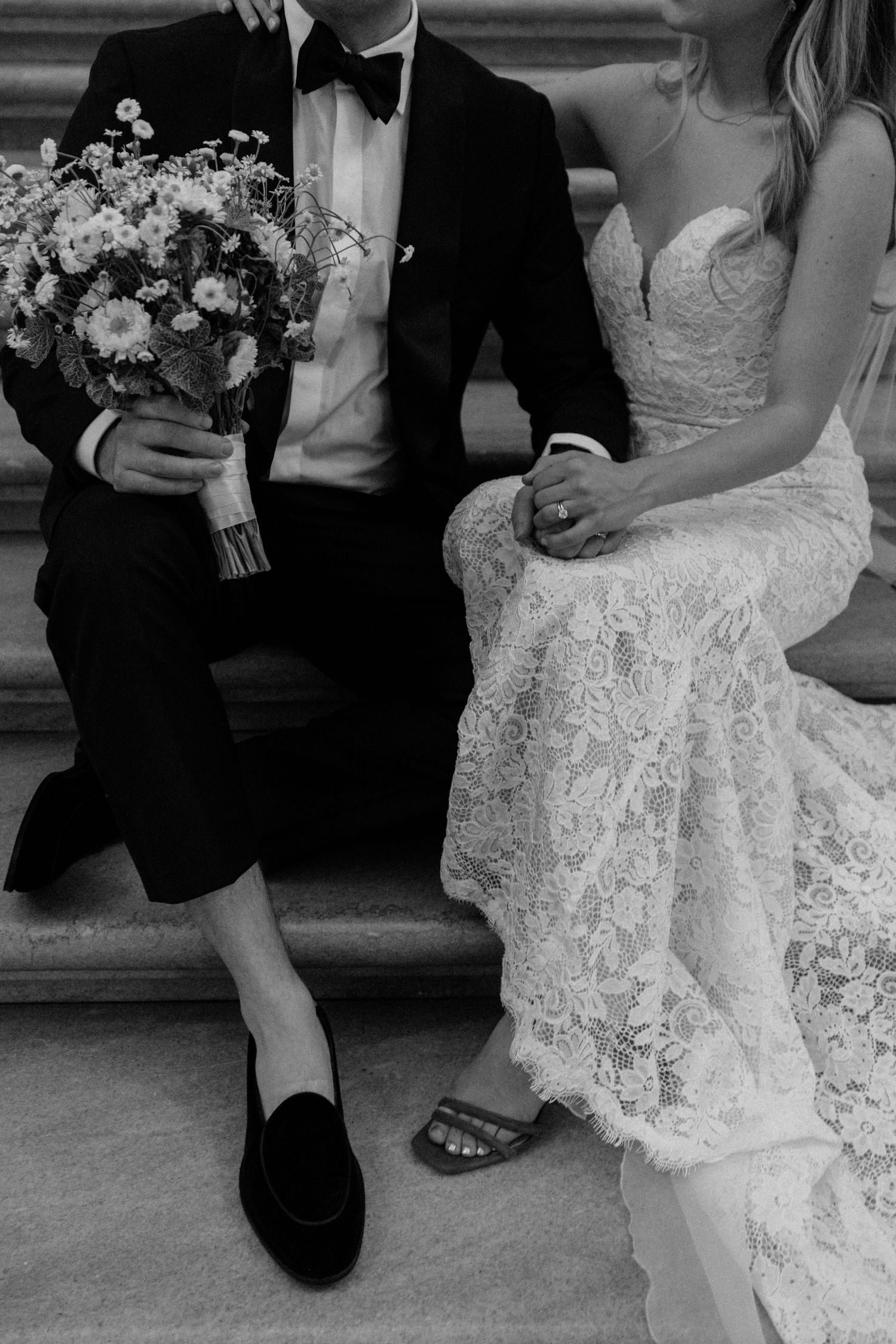 Cropped black and white photo showing wedding attire and bouquet detail with couple sitting on stone steps.