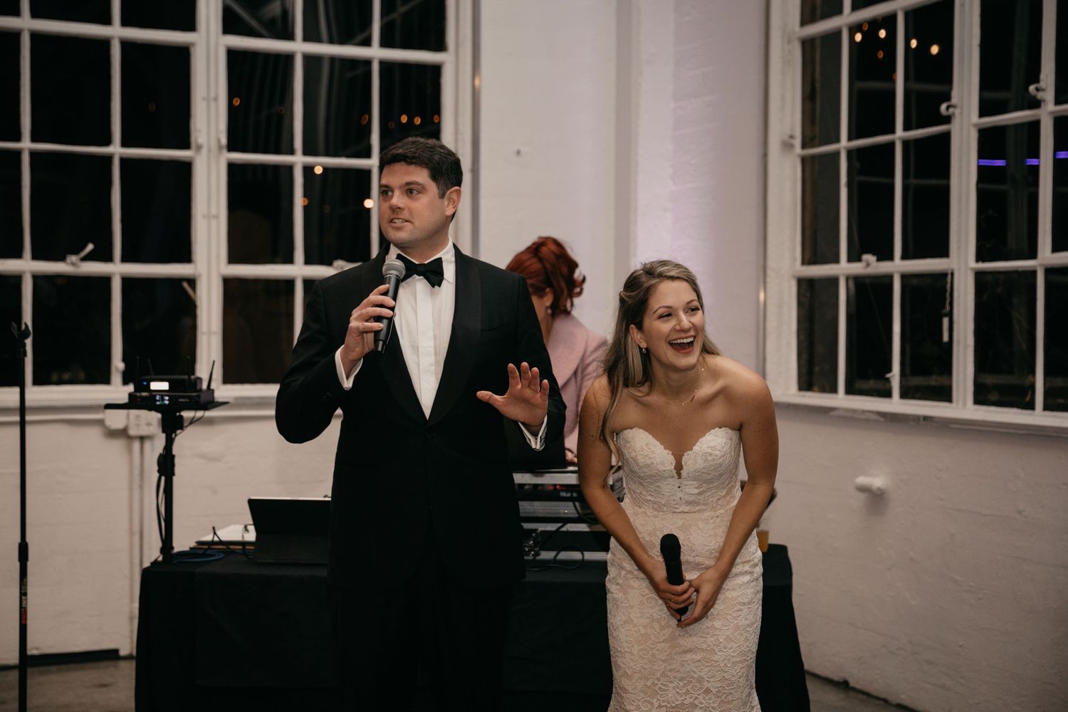 A couple gives speeches at their wedding reception in a bright white room with large windows.