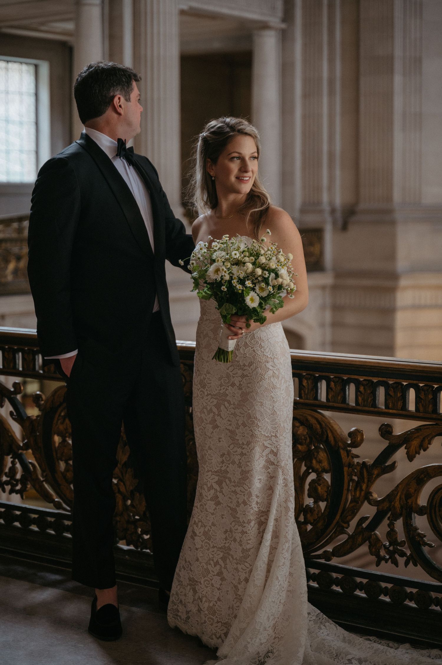 A wedding couple poses along an ornate railing in a grand marble venue with classic architectural details.
