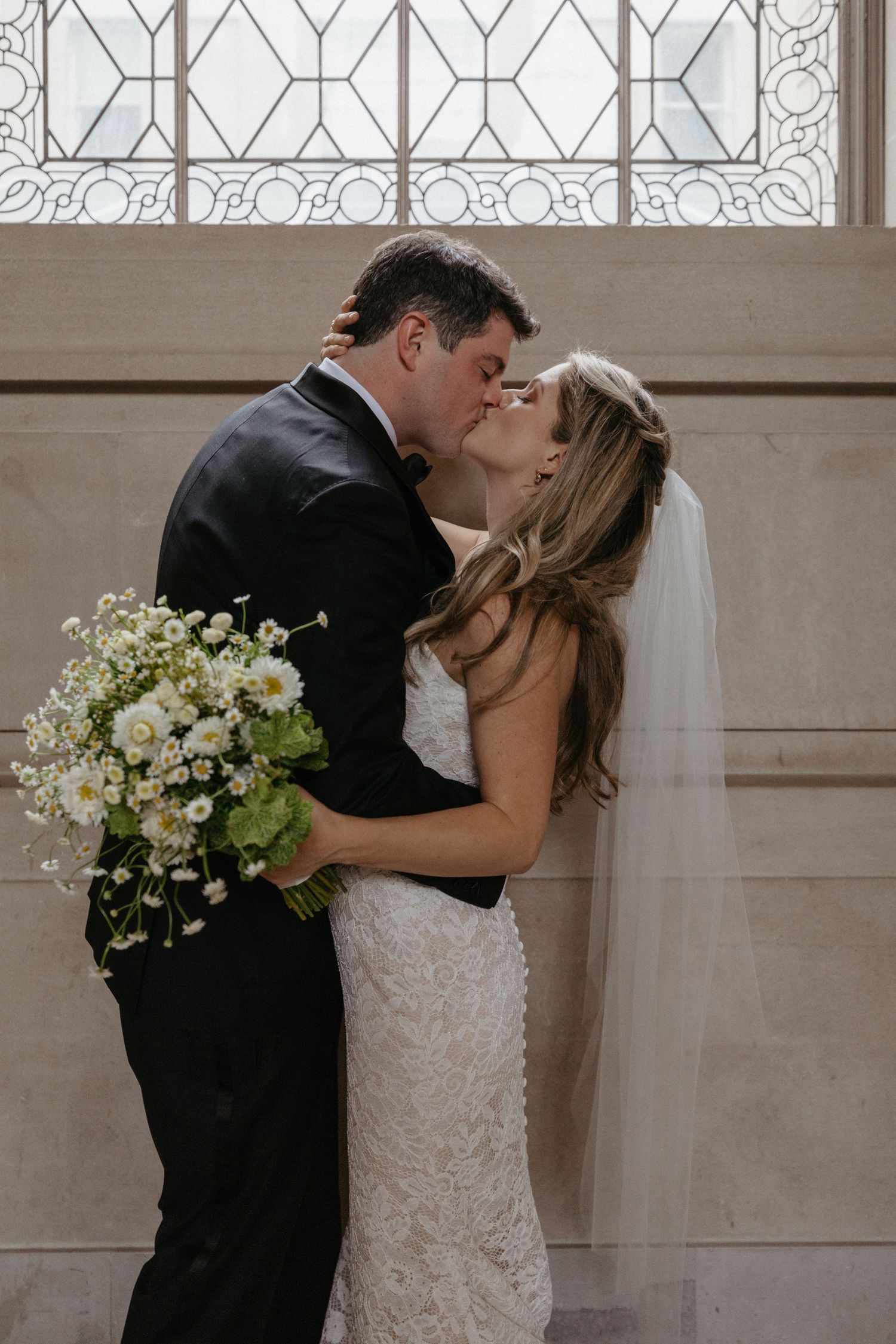 A romantic wedding moment captured near a decorative window as the couple shares a kiss with white flowers.