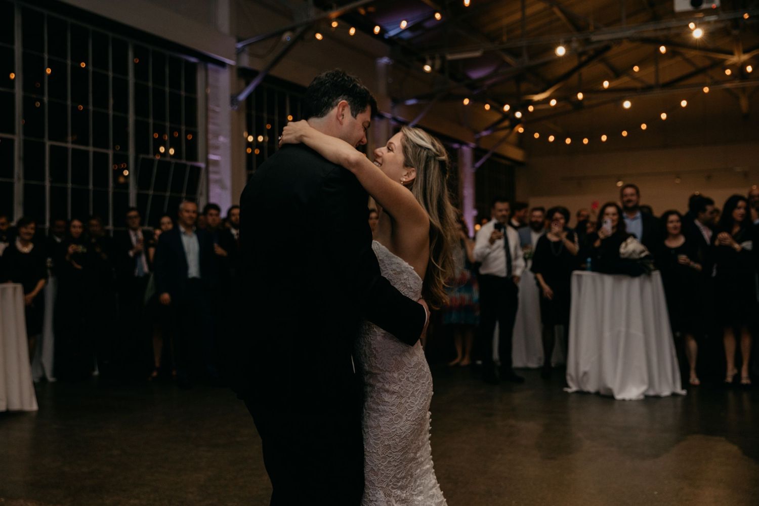 Couple sharing their first dance surrounded by guests at an indoor wedding reception.