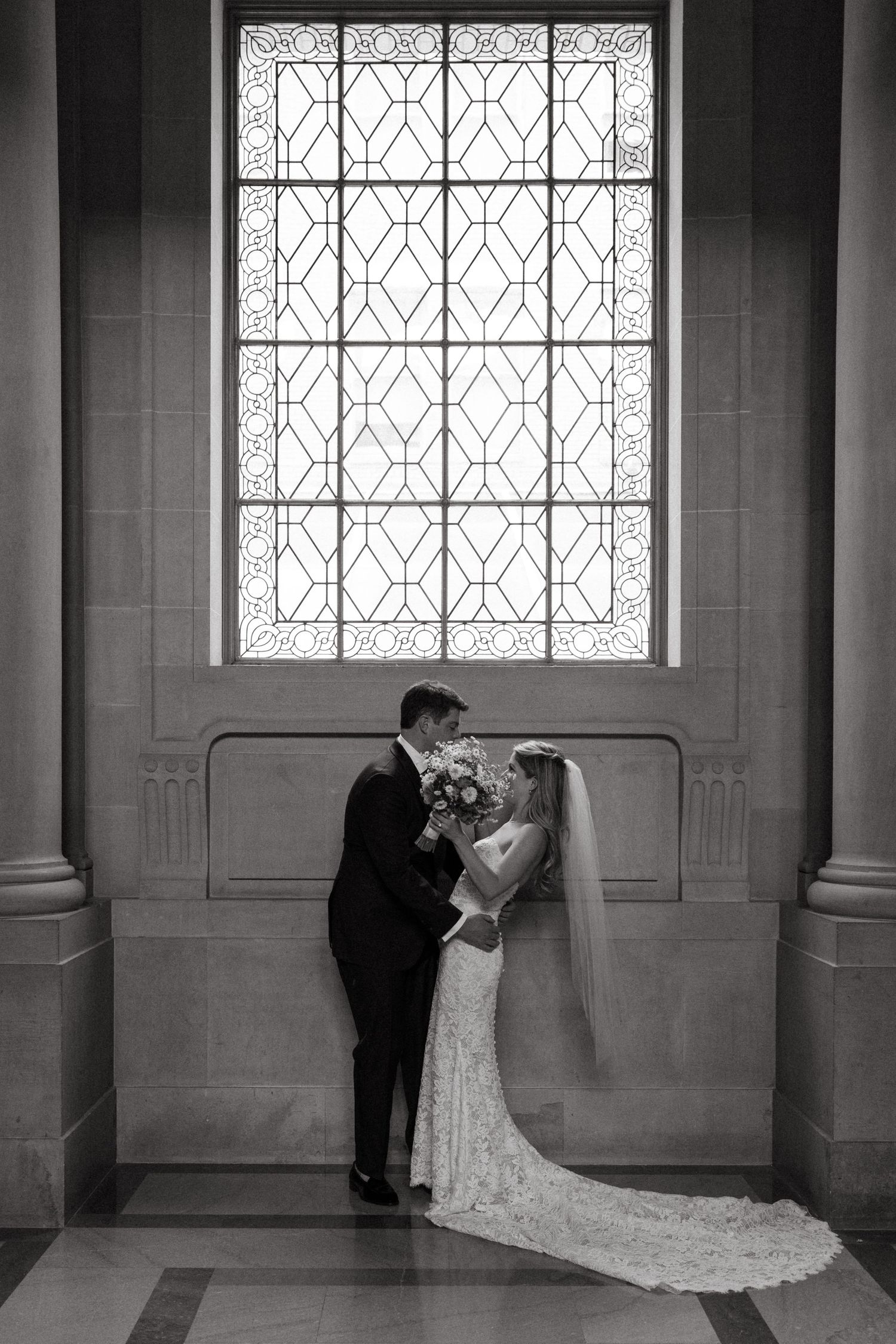 Black and white portrait of wedding couple beneath geometric window with flowing lace train.