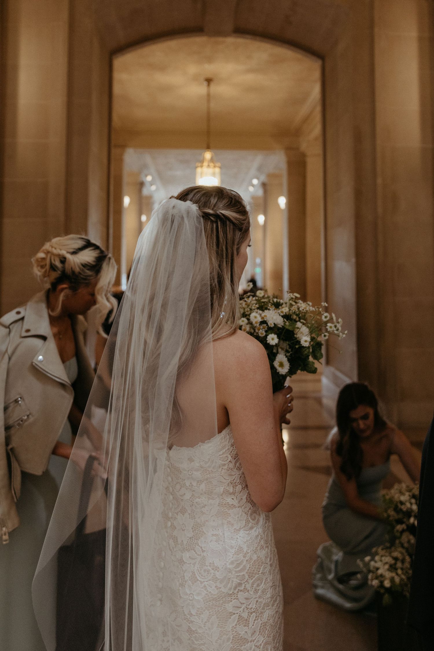 A bride in a lace wedding dress and long veil stands in an elegant hallway with ornate architectural details.