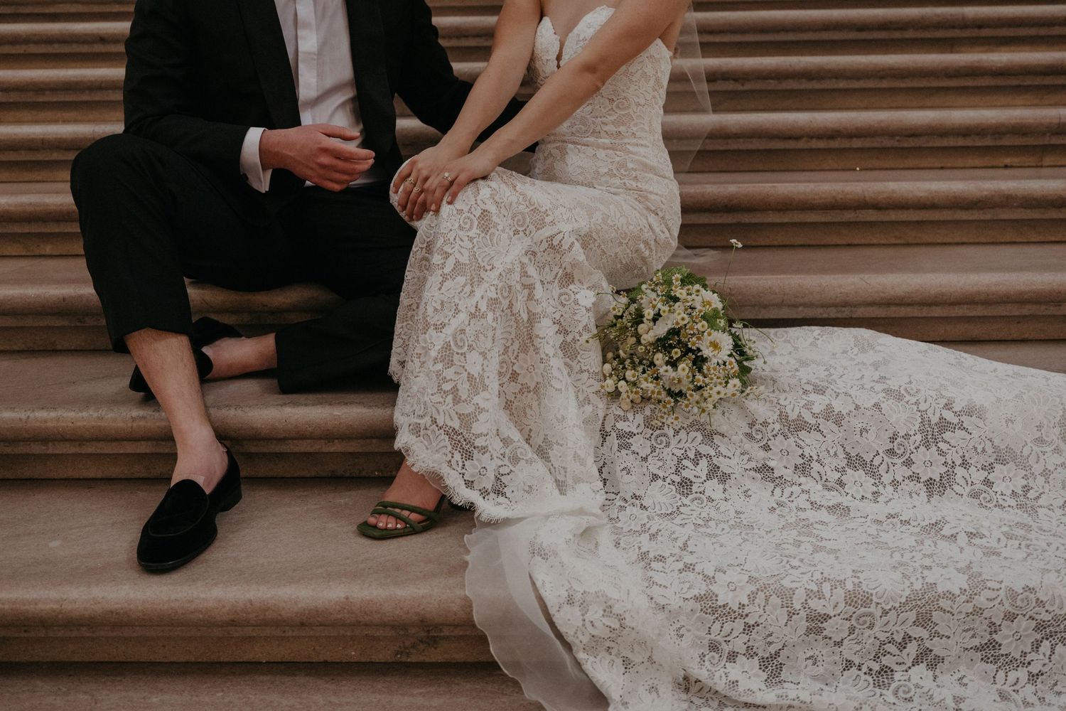 Detail shot of elegant lace wedding dress and black tuxedo as couple sits on stone steps.