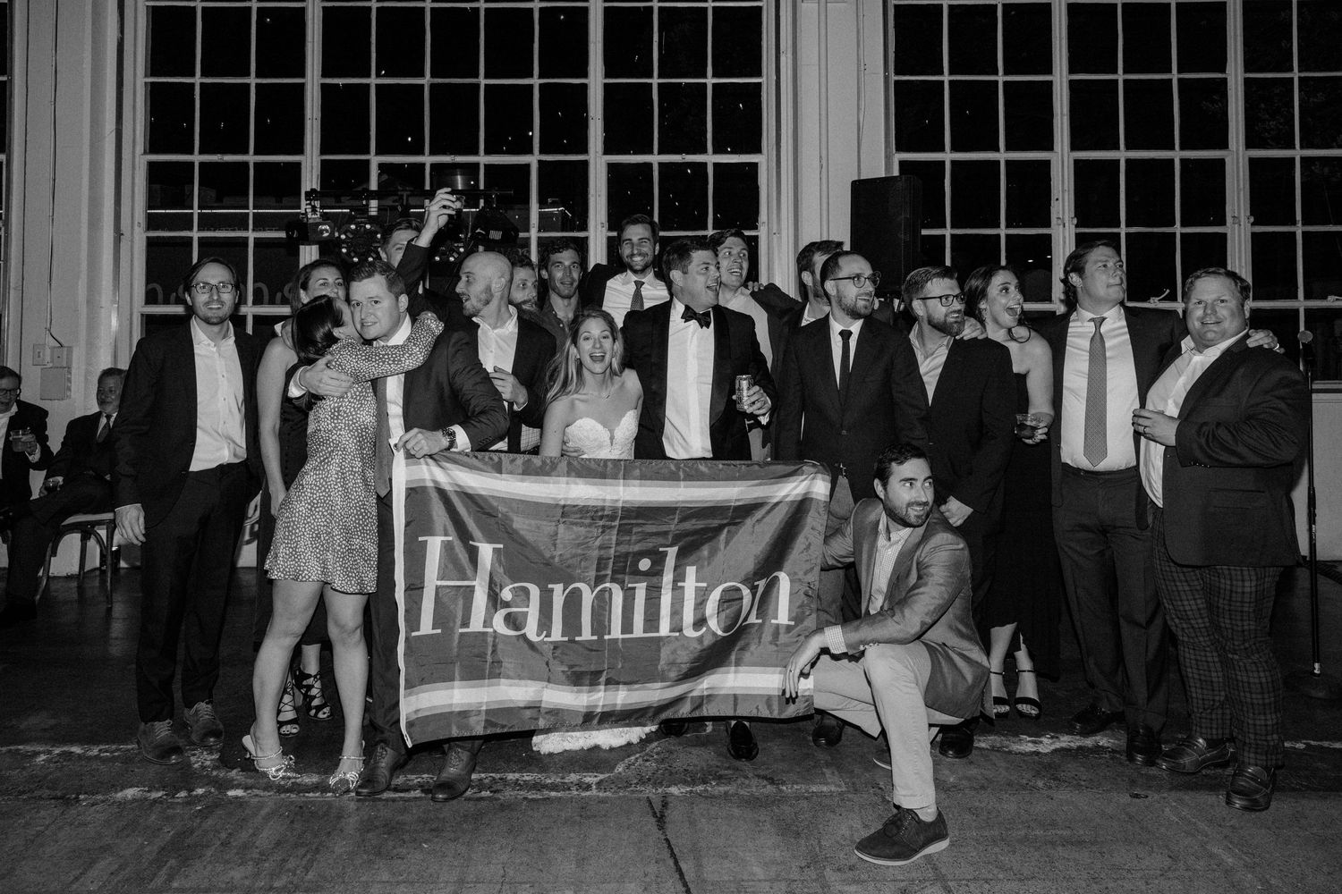 Group of wedding guests posing with a 'Families' banner in front of a windowed wall at night.