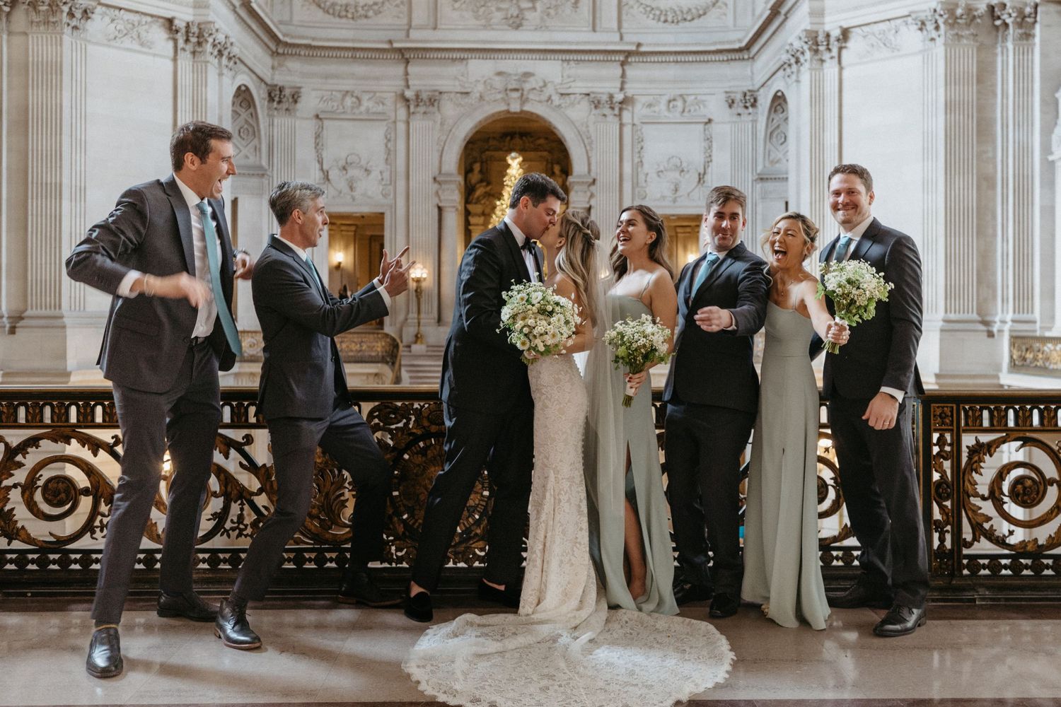 Wedding party poses for a fun group photo along ornate railings in classic marble venue.