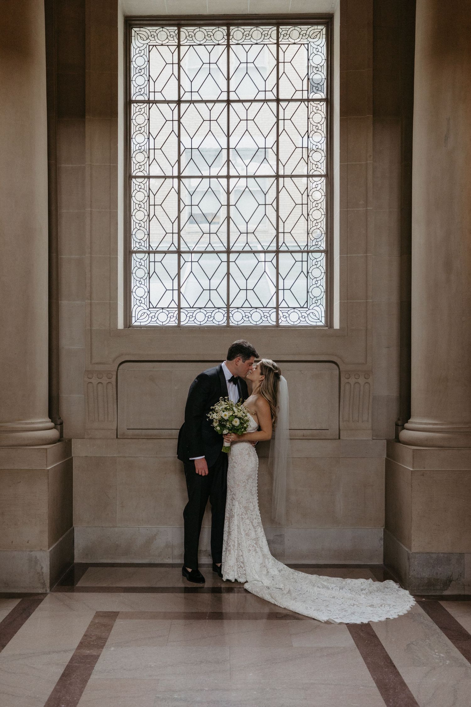 Wedding couple shares intimate moment beneath geometric window in classic architectural setting.