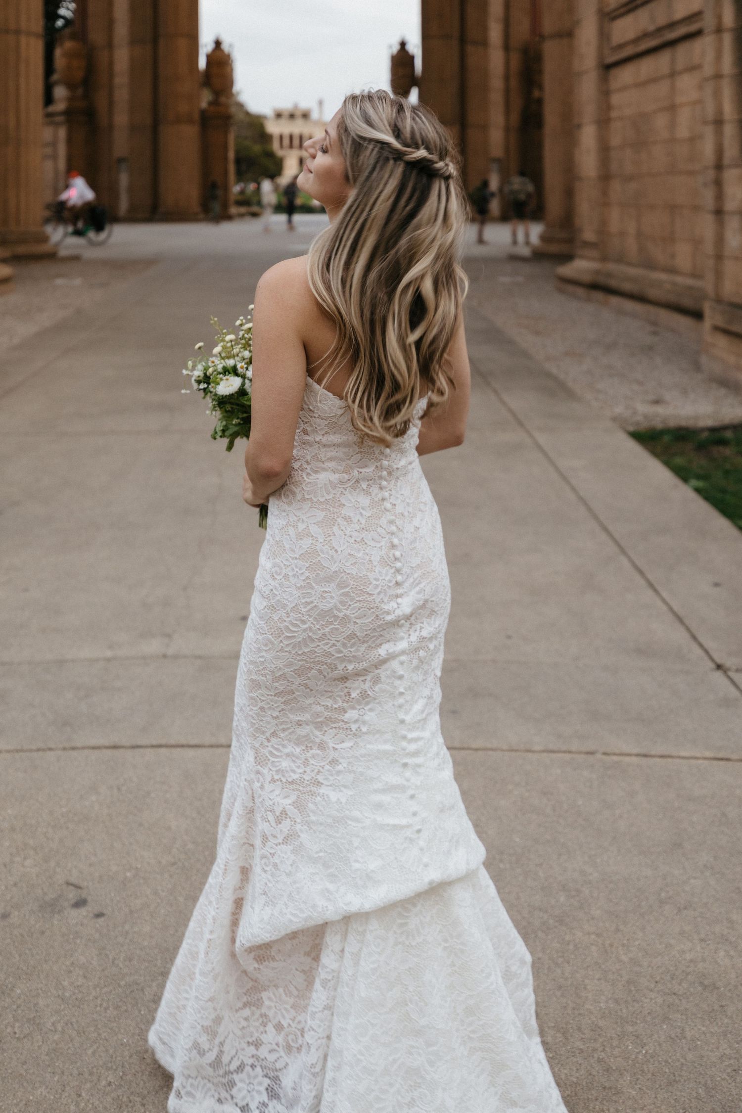 Back view of bride in fitted lace wedding gown holding bouquet between classical columns.