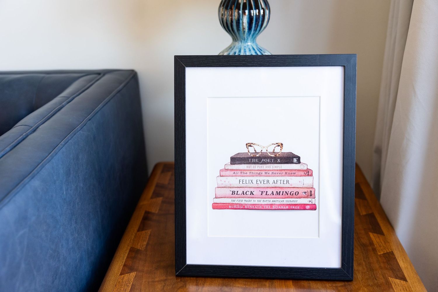 Framed print of stacked pink books on a wooden side table next to a blue sofa.