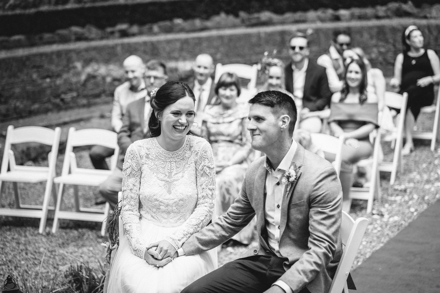 Black and white candid photo of newlyweds seated outdoors during wedding ceremony.