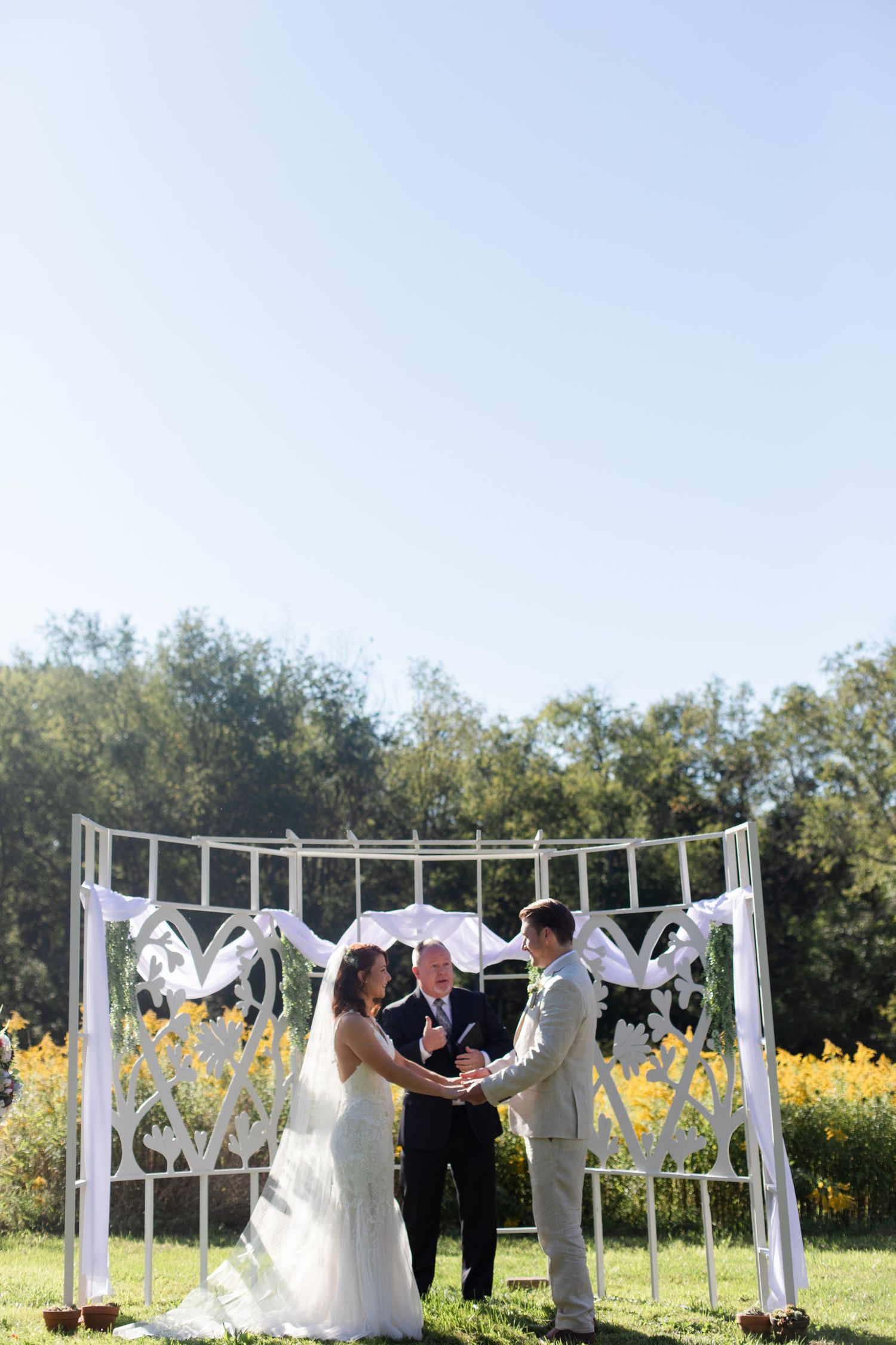 A wedding ceremony in progress under a white decorated arch with yellow flowers in the distance.