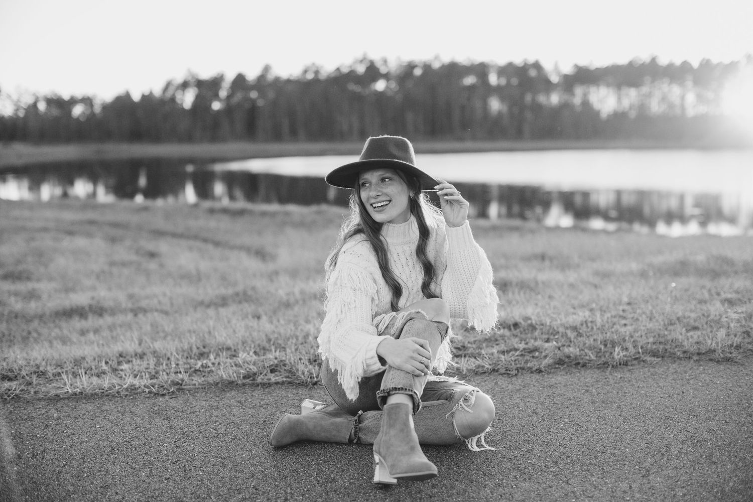 A black and white portrait of someone in a wide-brimmed hat crouching on a path near a lake.