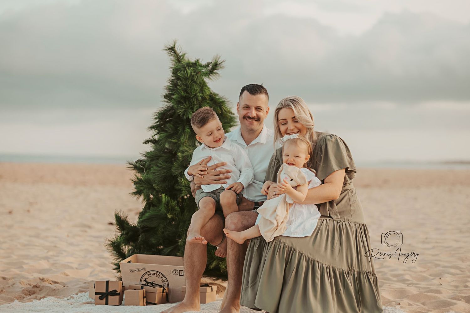 Family of four posing by a Christmas tree on the beach with holiday gifts.