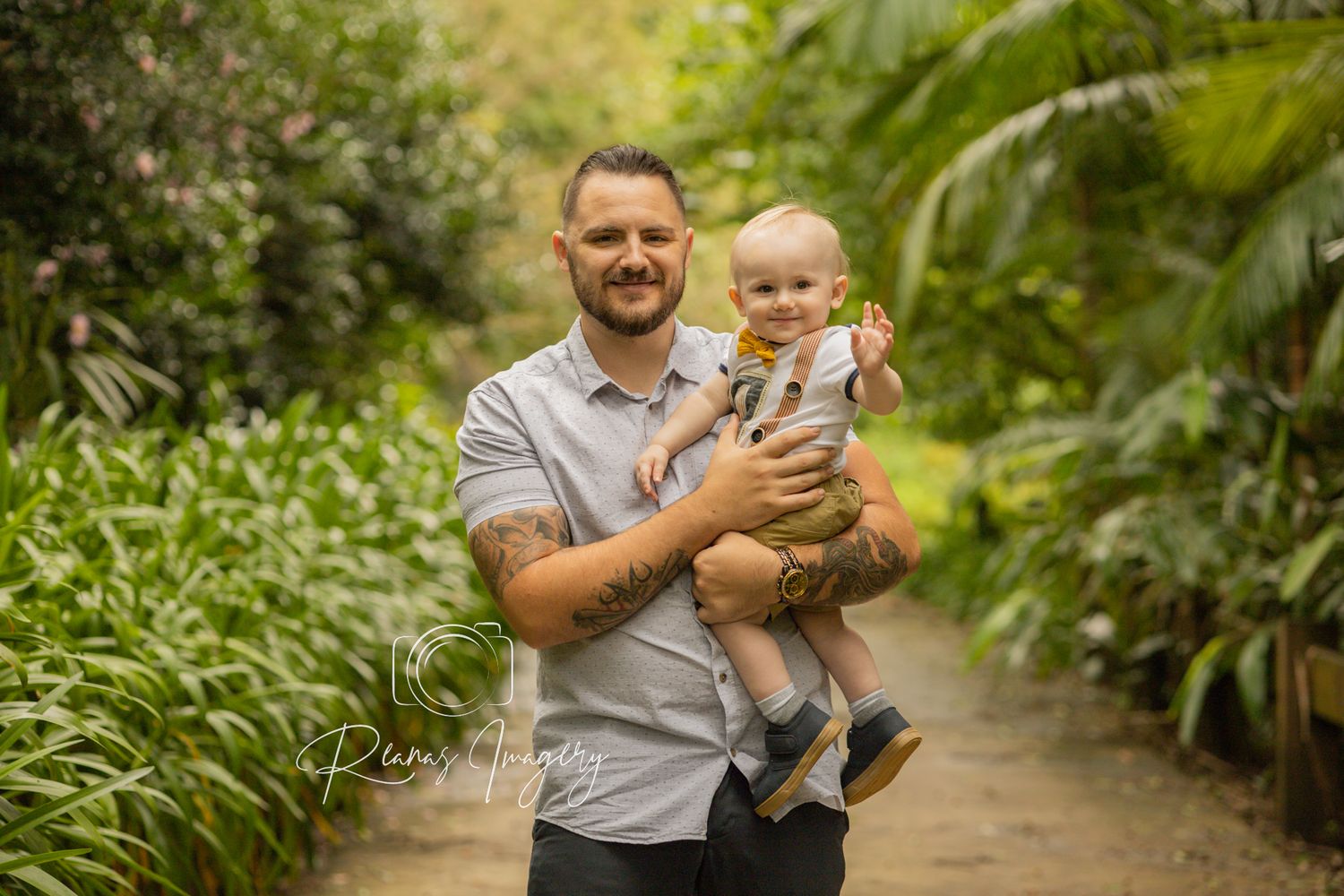 Smiling father with tattoos holding baby in lush green botanical garden path.