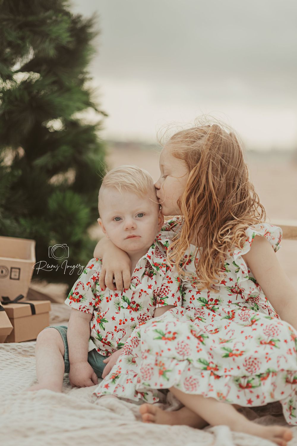 Young girl kissing baby brother wearing matching floral outfits near a Christmas tree.