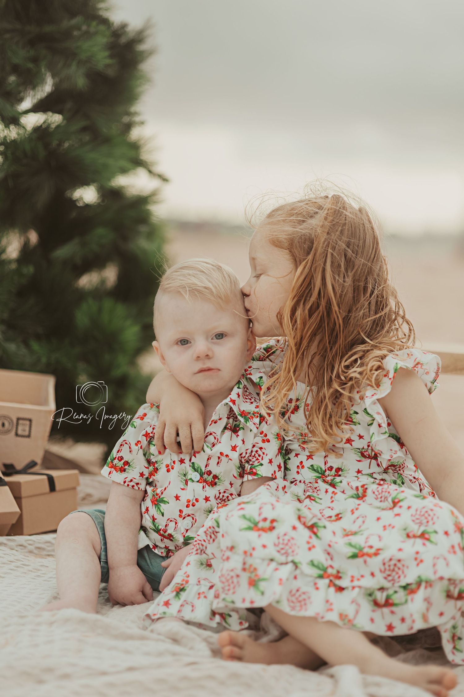 Young girl kissing baby brother wearing matching floral outfits near a Christmas tree.