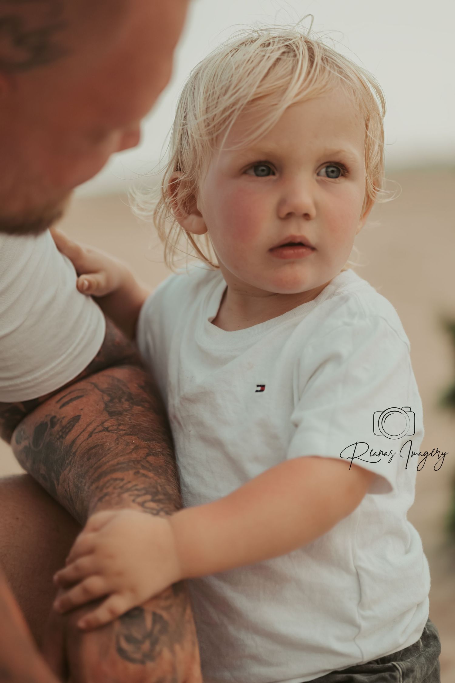 Blonde toddler boy in white Tommy Hilfiger shirt held by tattooed father at beach.