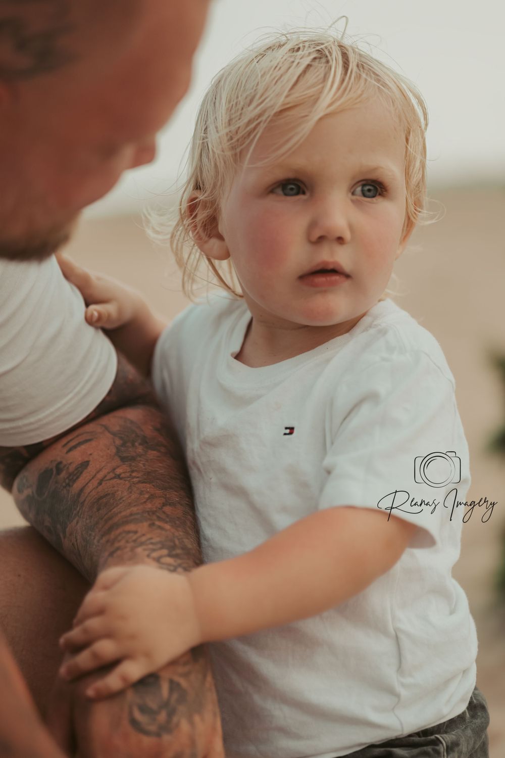 Blonde toddler boy in white Tommy Hilfiger shirt held by tattooed father at beach.