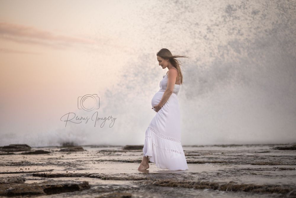 Pregnant woman in flowing white dress standing on rocky shore as a dramatic wave crashes behind her.
