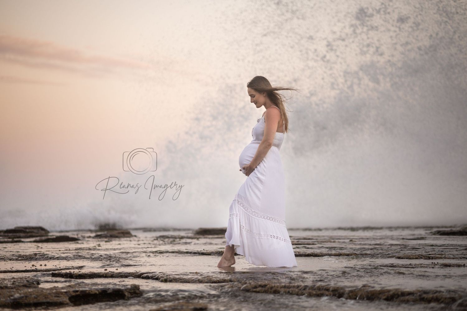 Pregnant woman in flowing white dress standing on rocky shore as a dramatic wave crashes behind her.
