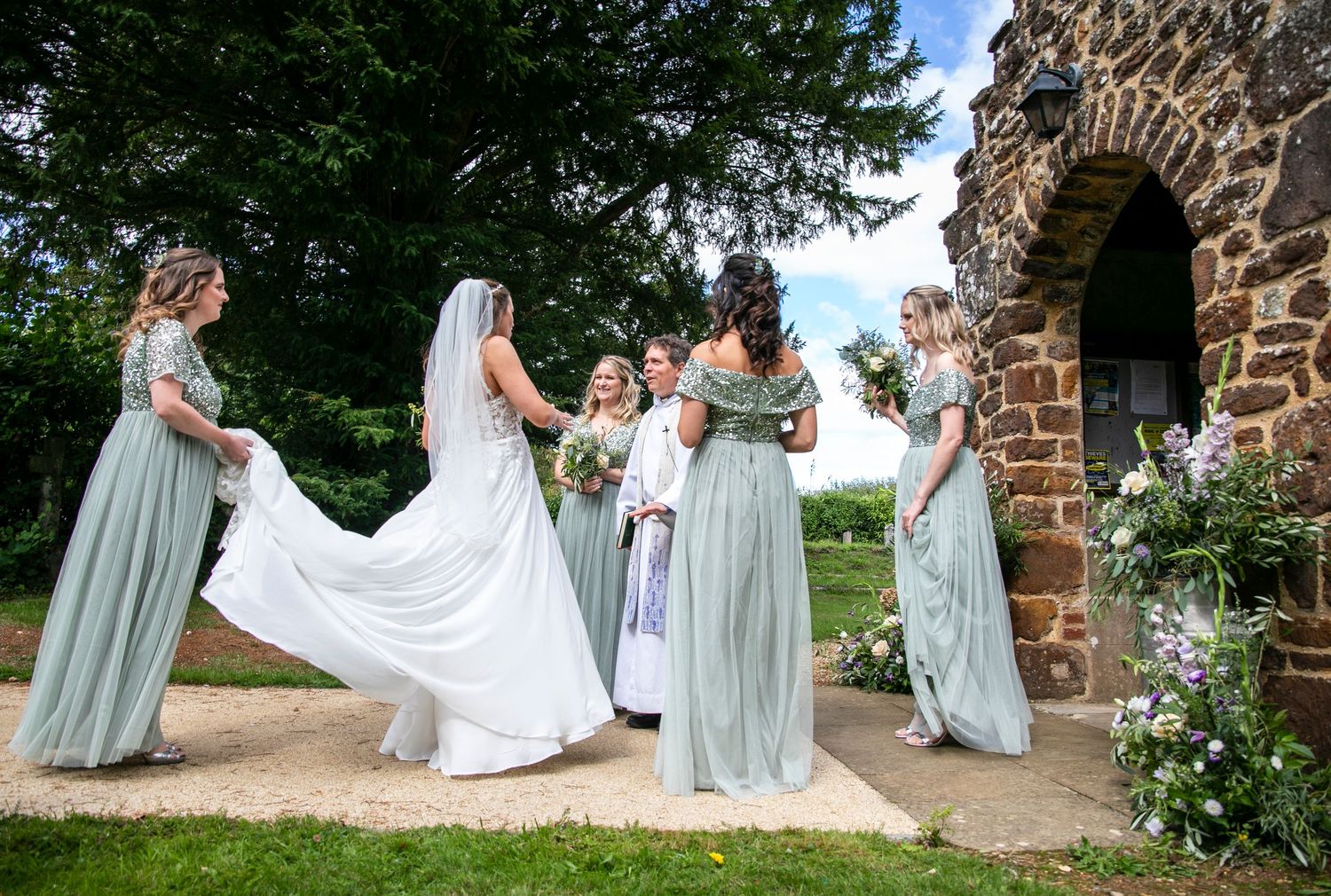 Wedding party poses in front of stone wall with bridesmaids in sage green dresses.