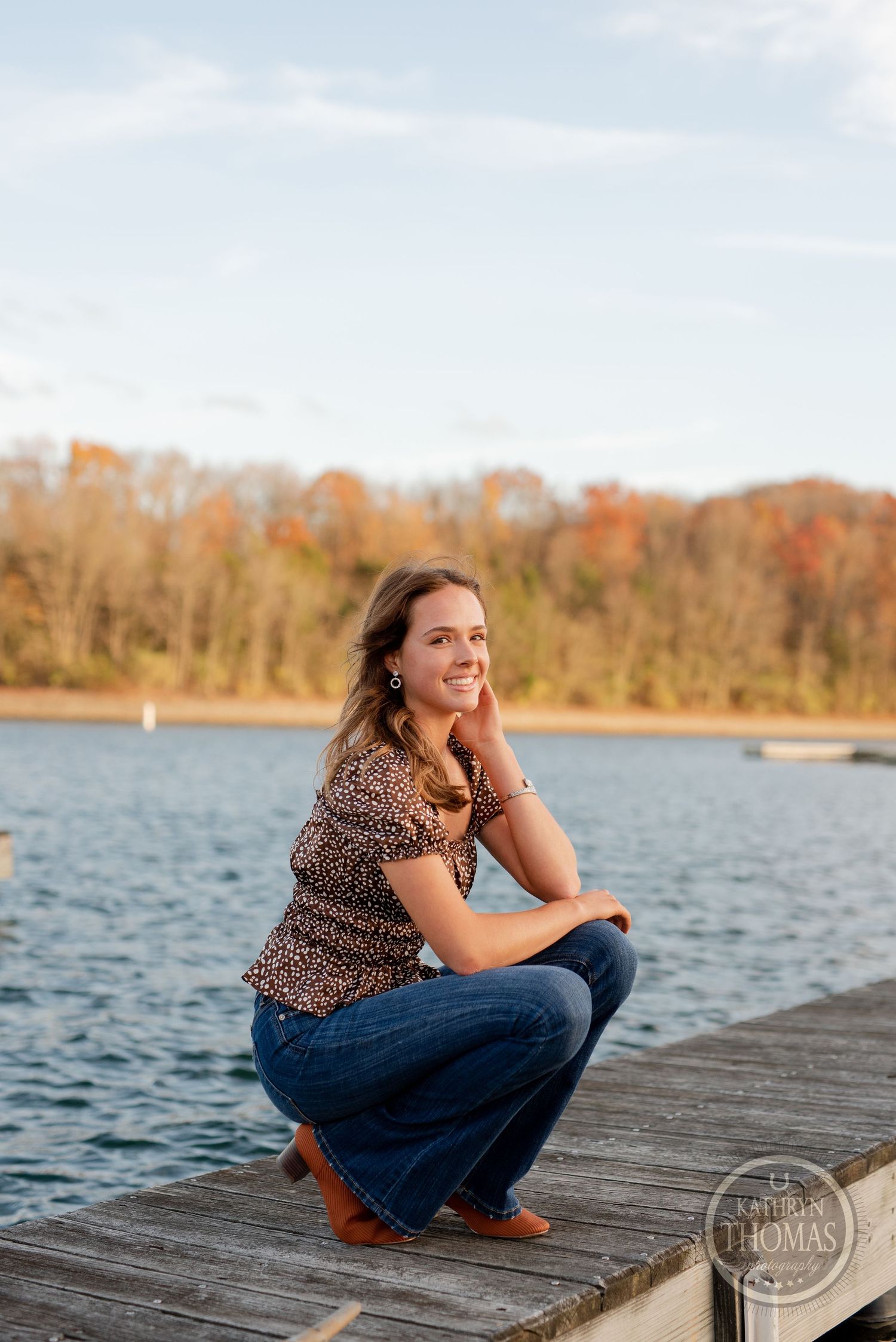 Someone crouching on a wooden dock by the lake during sunset with fall foliage in the background.
