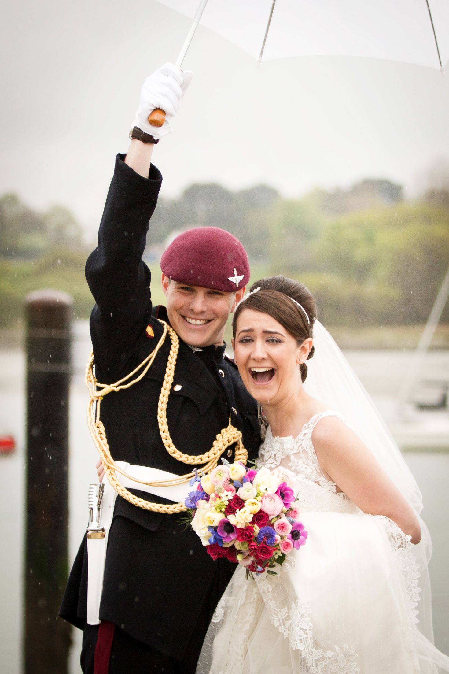 Joyful wedding couple celebrates in the rain with white umbrella by waterfront.