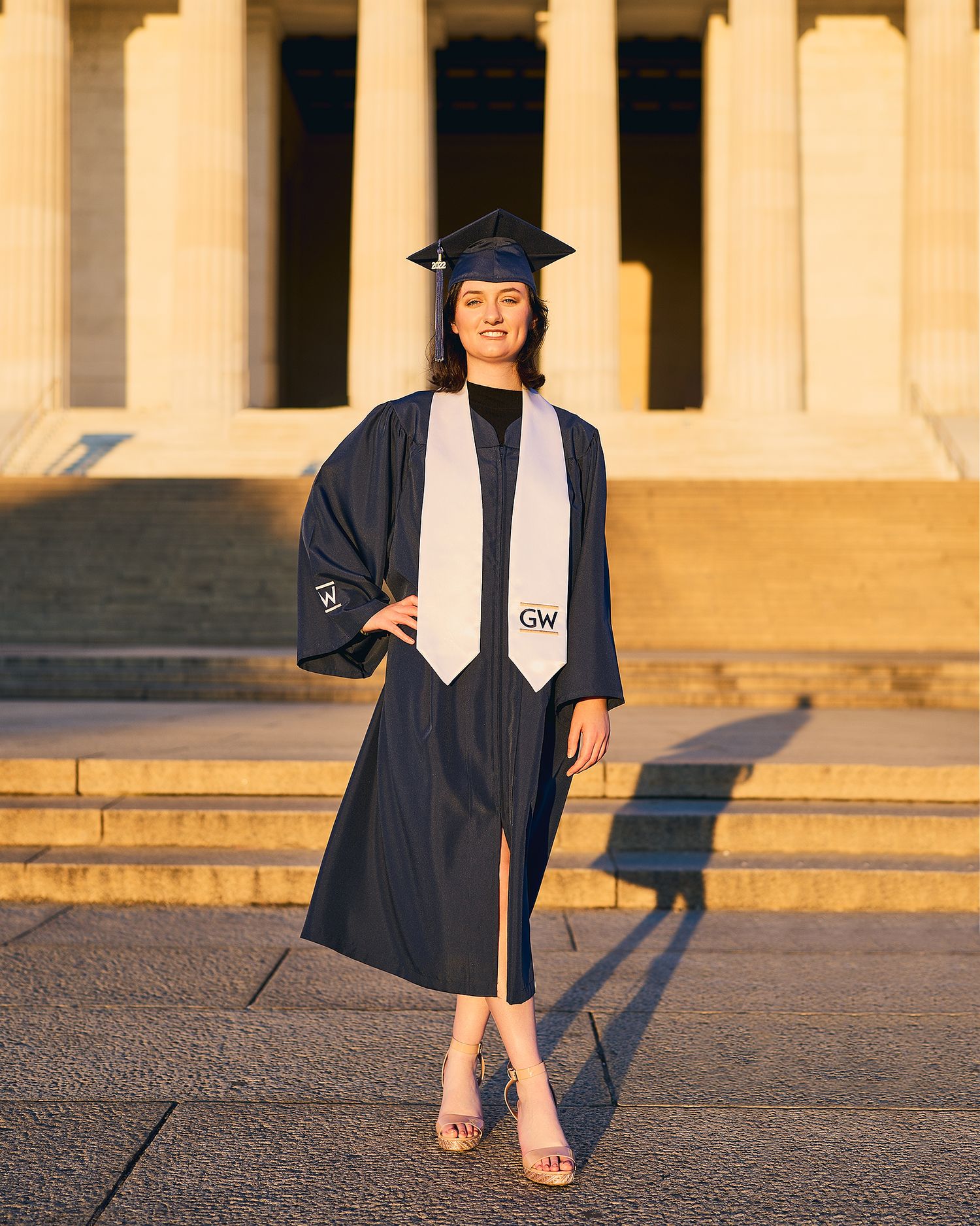 Graduate in traditional cap and gown poses on sunny steps of academic building.