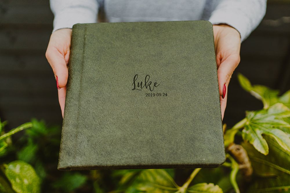 Hands holding an olive green velvet-covered book against foliage background.