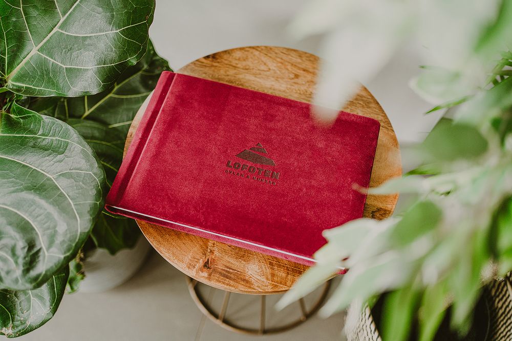 Red velvet book lying on wooden surface surrounded by green plants.