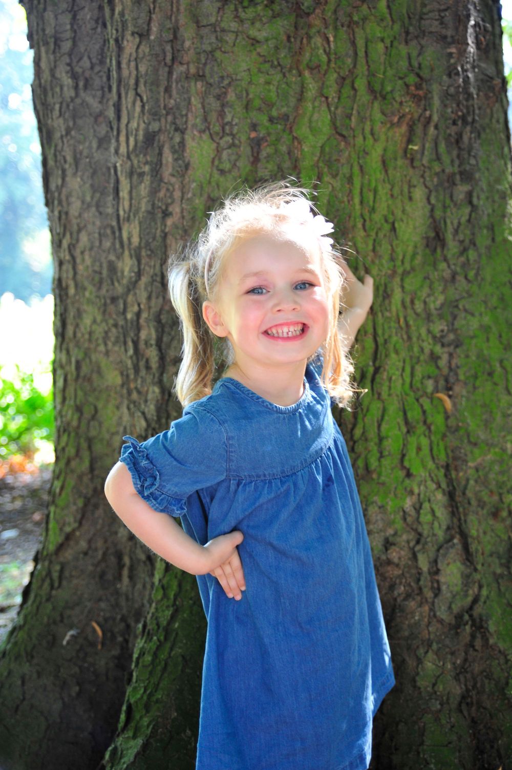 A small child in a blue denim dress poses playfully against a large tree trunk with moss.