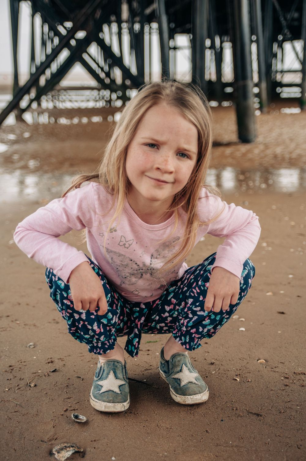 A casual portrait taken under a pier with someone crouching on sandy beach.