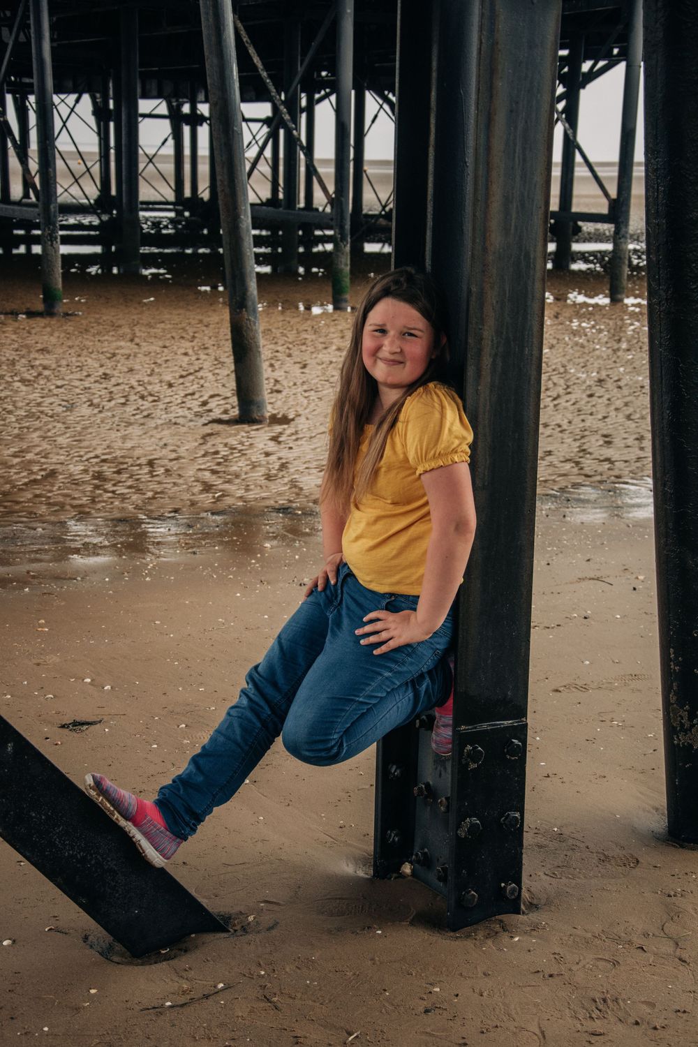 A casual portrait under a pier featuring yellow shirt and jeans against beach backdrop.