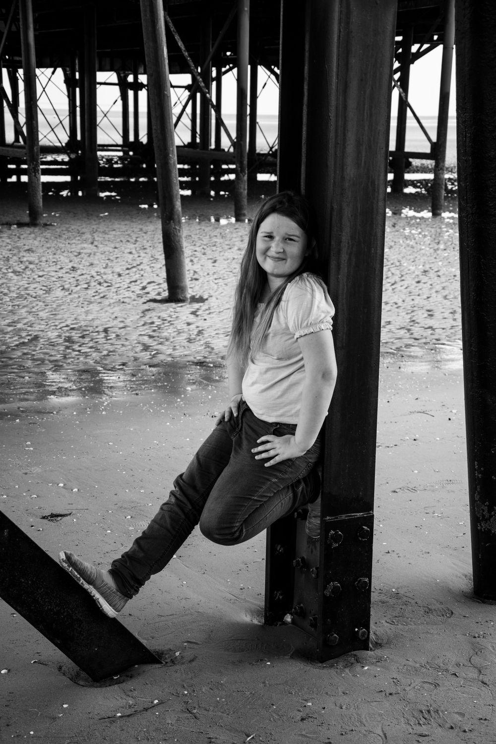 A black and white photo showing someone posing playfully between pier supports.