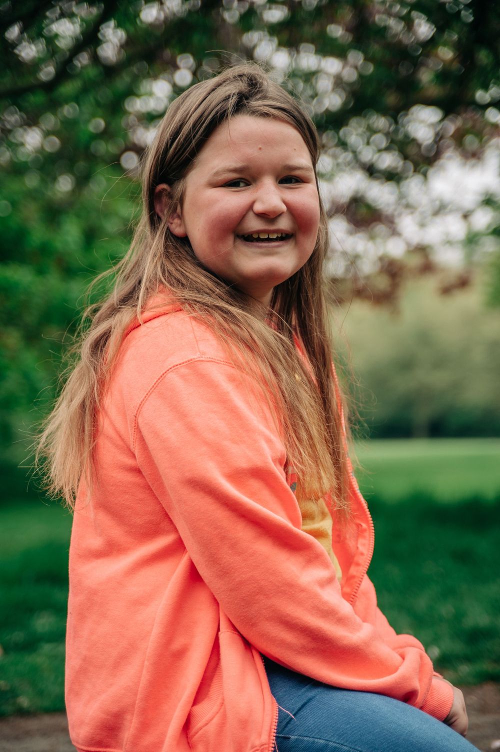A person in a coral sweatshirt smiles brightly in a green park setting.