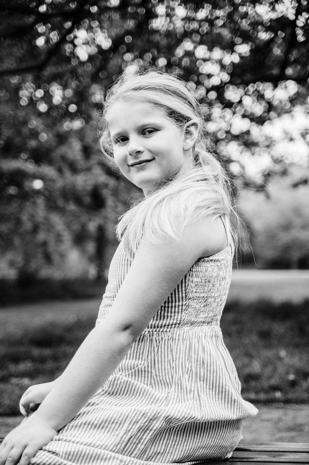 A black and white portrait of a child in a striped dress sitting outdoors with bokeh background.