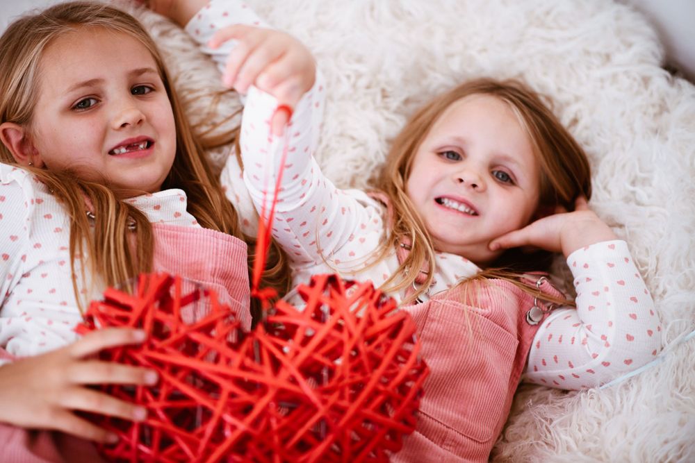 Two young children lying on white bedding playing with large red heart decoration.