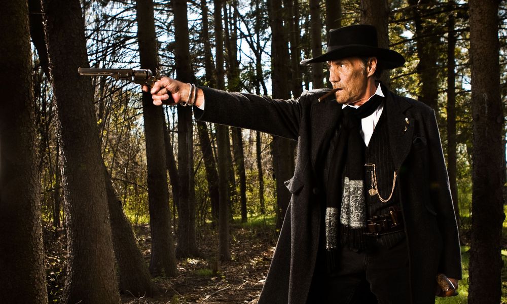 Cowboy in a top hat and coat holds a gun while standing in the woods.