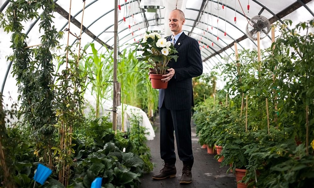 A well-dressed man holding a plant in a greenhouse, showcasing the perfect blend of sophistication and nature's beauty.