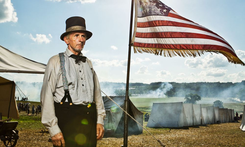 Senior gentleman in formal attire poses near patriotic tent with US flag.
