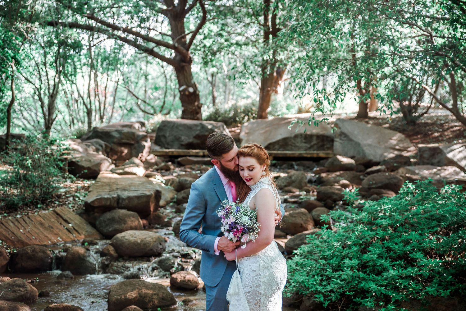 Couple embracing by rocky stream in woodlands during wedding photoshoot.
