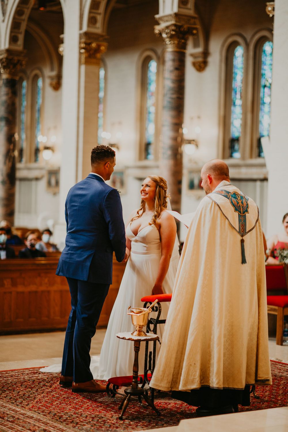 Smiling bride in white gown and groom in blue suit exchange vows with officiant.