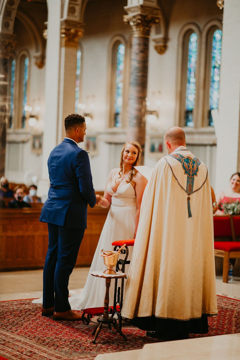 Bride and groom holding hands during wedding vows with officiant in ornate church.