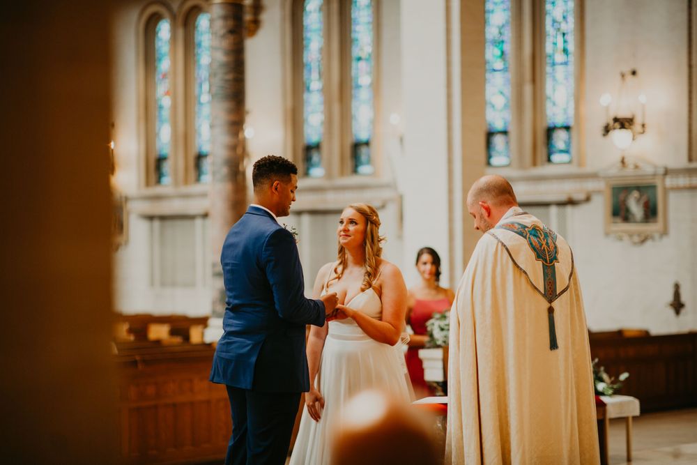 Couple holds hands before officiant during elegant church wedding ceremony with stained glass windows.