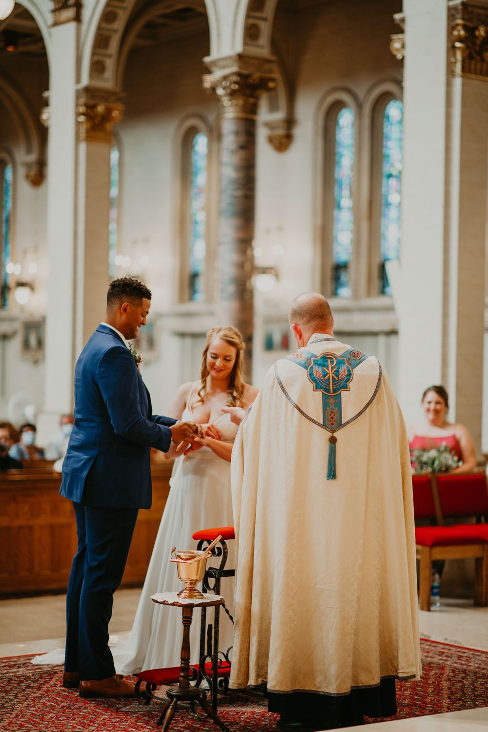 Groom places wedding ring on bride's finger as priest in decorative vestments officiates church ceremony.