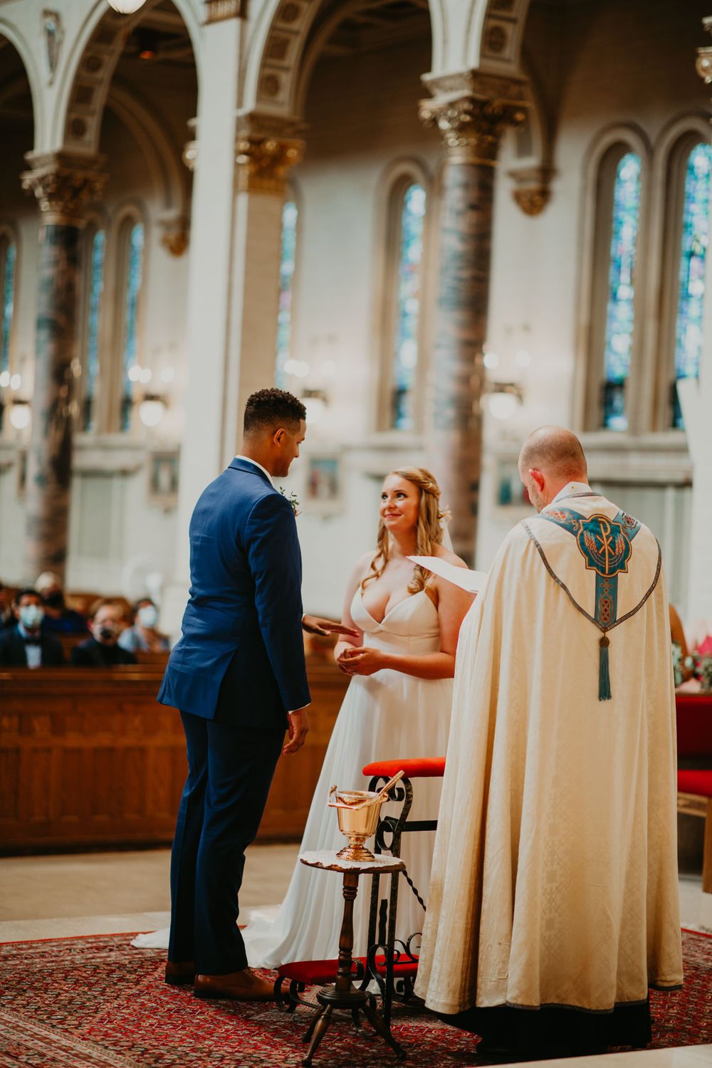 Smiling bride receives wedding ring from groom during formal church ceremony with priest in embroidered robe.